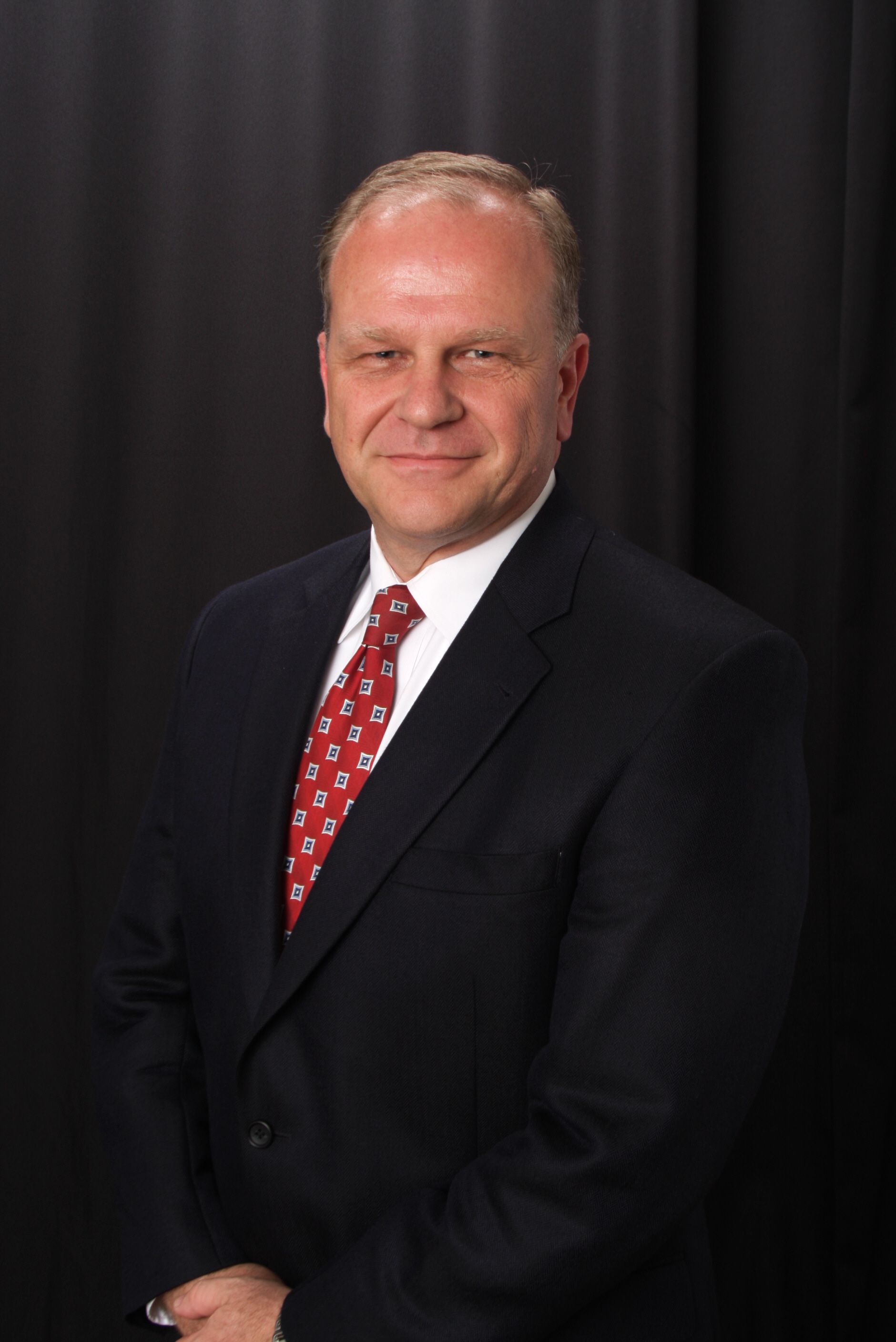 Man in a dark suit with a red patterned tie, smiling. Standing in front of a dark curtain. Man in a dark suit with a red patterned tie, smiling. Standing in front of a dark curtain.