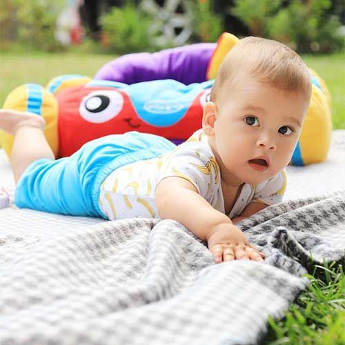 A baby lying on their stomach on a blanket outdoors, next to a colorful plush toy with a cartoon face.