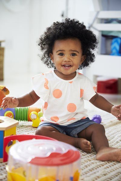 A smiling toddler with curly dark hair sits on a rug surrounded by colorful plastic toys.
