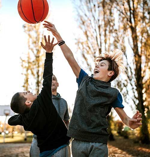 Two people reach toward a basketball in mid-air on an outdoor court, while a third person watches in the background.