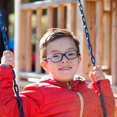 A person wearing blue glasses and a red jacket smiles while holding onto blue swing chains on a playground.