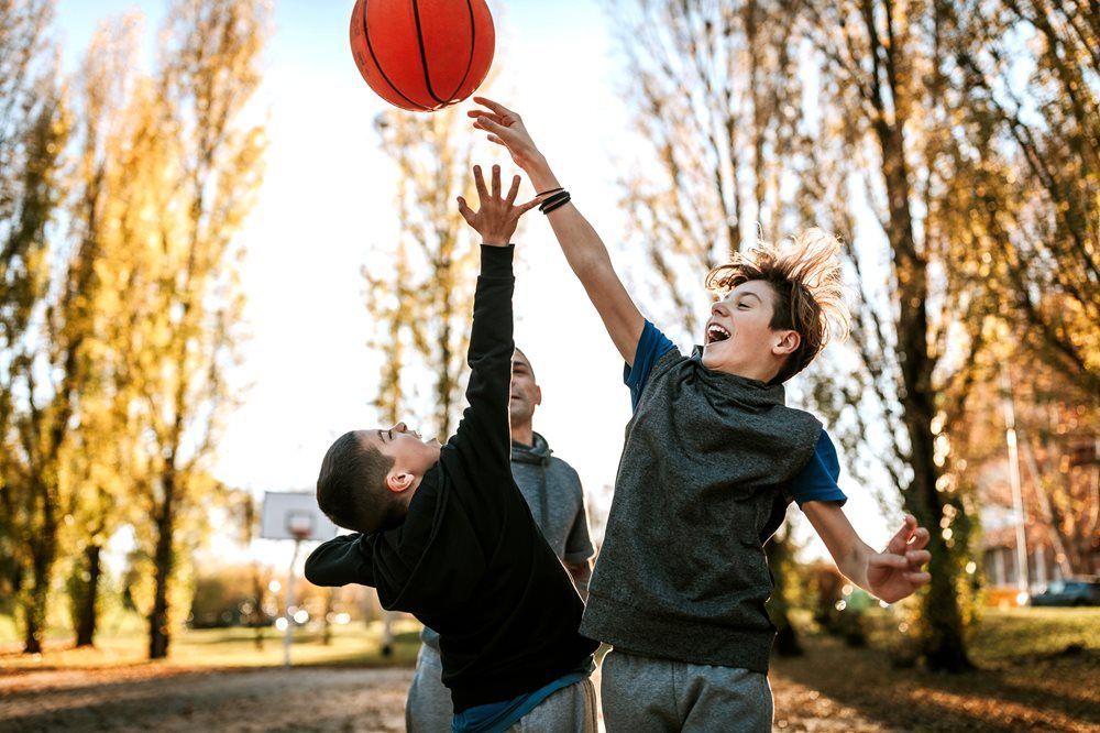 Two people jump to reach a basketball on an outdoor court surrounded by trees during autumn.