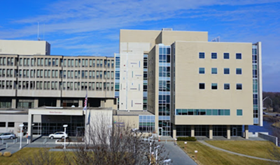 A large, tan hospital building with multiple stories, many windows, and a covered main entrance on a clear, sunny day.
