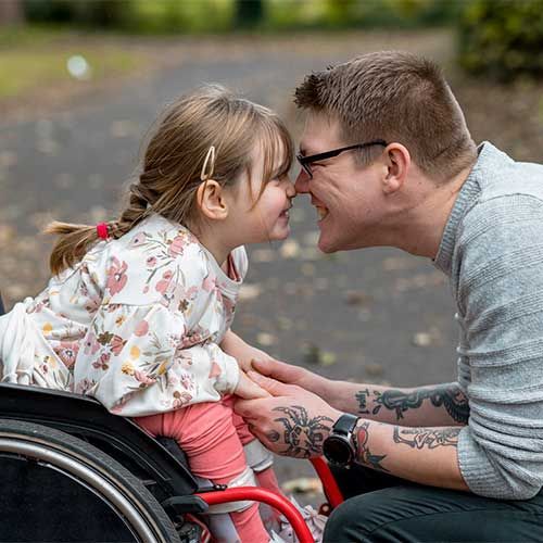 A person in a wheelchair and an adult touch foreheads, smiling at each other in a park setting.