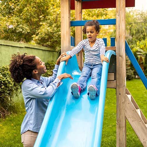 A person in a light blue shirt smiles and watches a child sitting at the top of a bright blue playground slide.