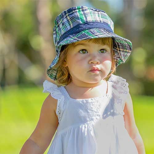 A toddler wears a green, blue, and white plaid bucket hat and a white dress outdoors in a softly blurred park.