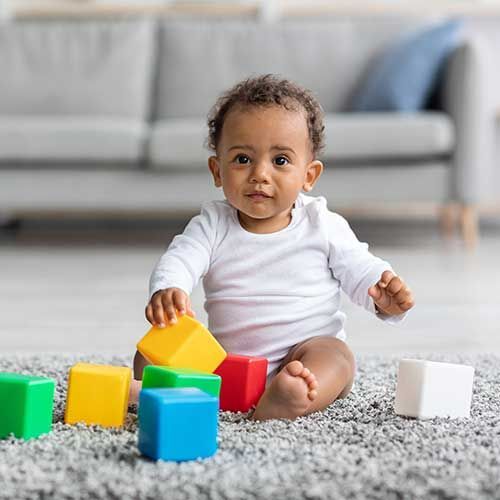 A baby in a white long-sleeve shirt sits on a grey rug, playing with colorful blocks in front of a sofa.