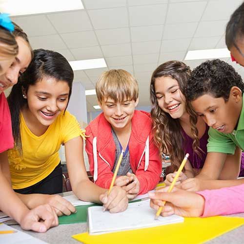 A group of students smiling and collaborating around a table while writing in a notebook in a classroom.