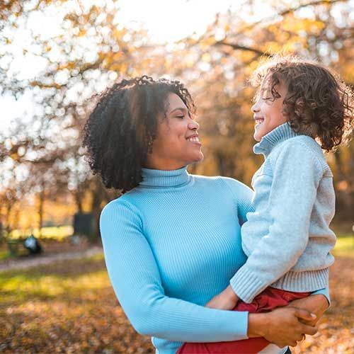 A person in a light blue turtleneck sweater holding a child in a matching sweater, smiling together in a sunny park.