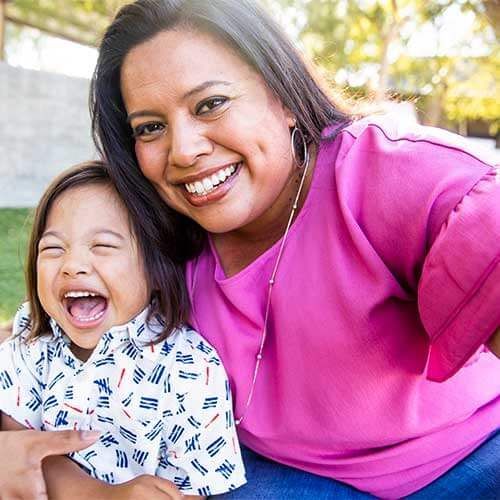 A smiling person holds a laughing child in a bright outdoor setting, both wearing casual, colorful clothing.