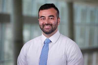 A man with a short beard smiles, wearing a white collared shirt and a blue patterned tie against a blurred office background.
