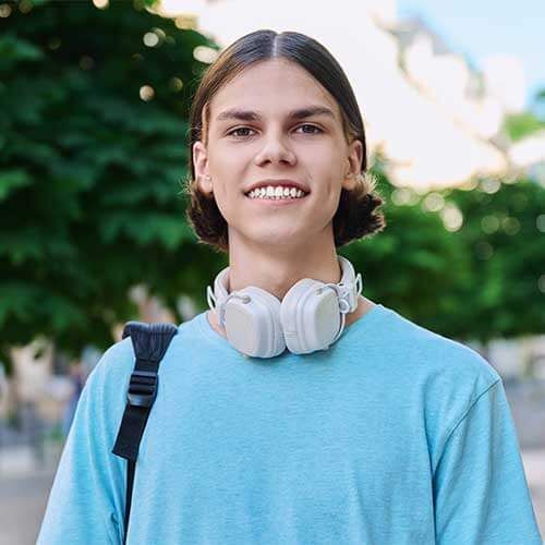 A person with shoulder-length brown hair and white headphones around their neck smiles while wearing a blue shirt.