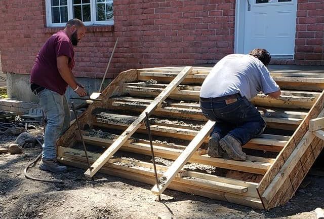 Deux hommes construisent des escaliers en bois devant un bâtiment en briques. L'un prend des mesures, l'autre travaille sur la structure.