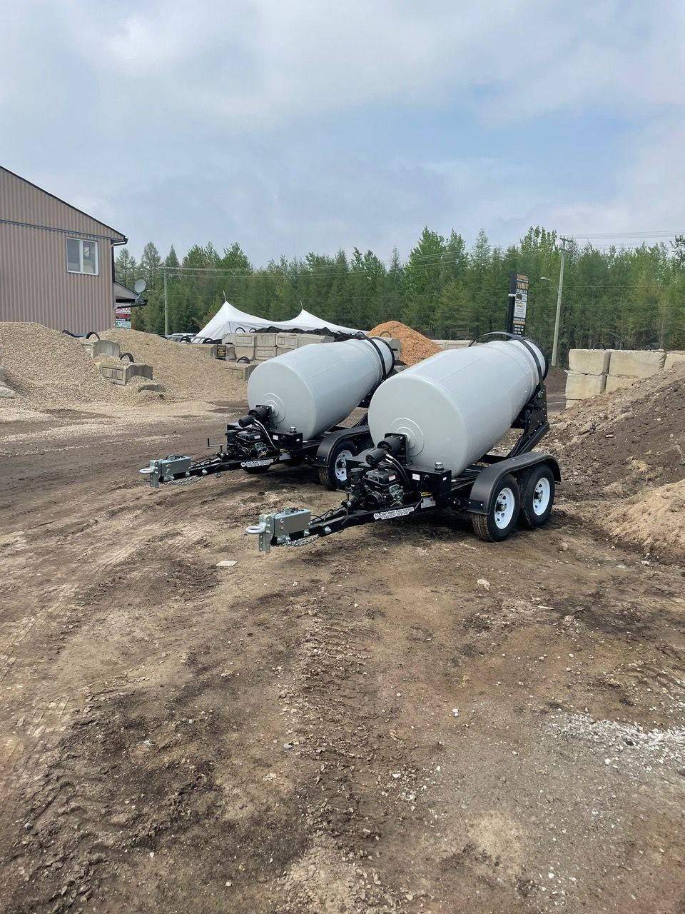 Deux remorques-citernes grises sur un terrain en terre, près d'un bâtiment et d'arbres.