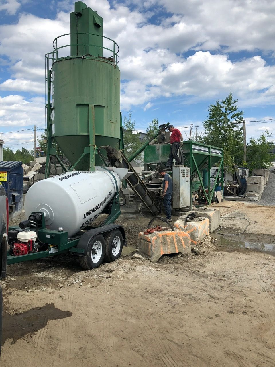 Remplissage d'une bétonnière sur un chantier. Des ouvriers se tiennent près des machines. Équipement vert et gris.