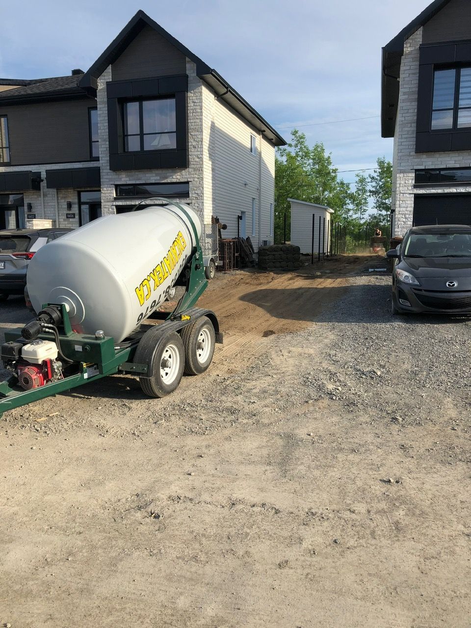 Bétonnière garée sur une allée de gravier près de maisons à deux étages sous un ciel nuageux.