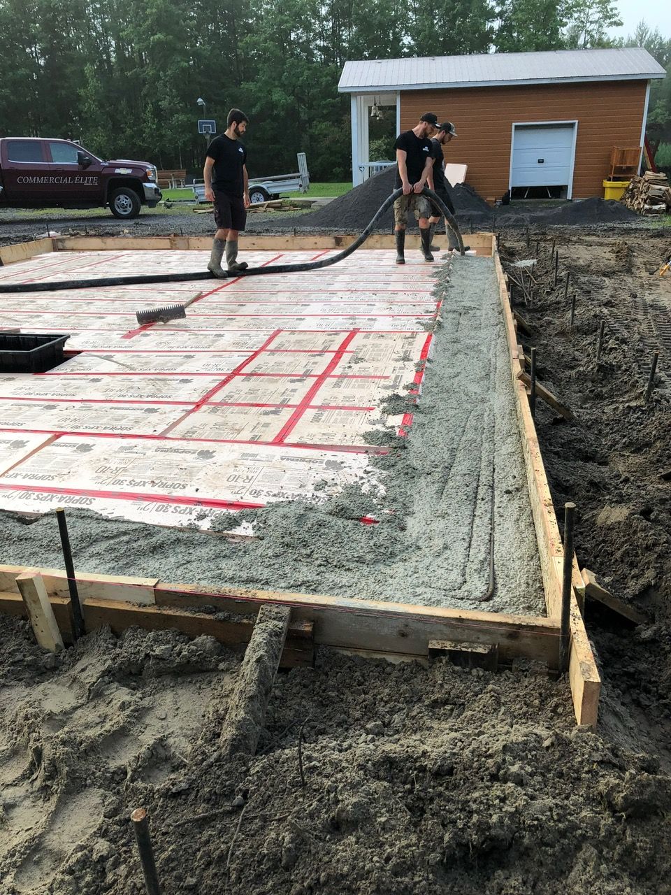 Trois ouvriers coulent du béton dans des fondations. Chantier extérieur avec un bâtiment en arrière-plan.