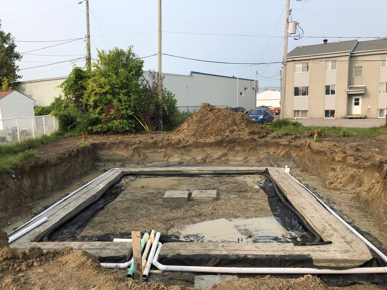 Chantier de construction avec cadre de fondation rectangulaire, entouré de terre excavée; canalisations visibles.