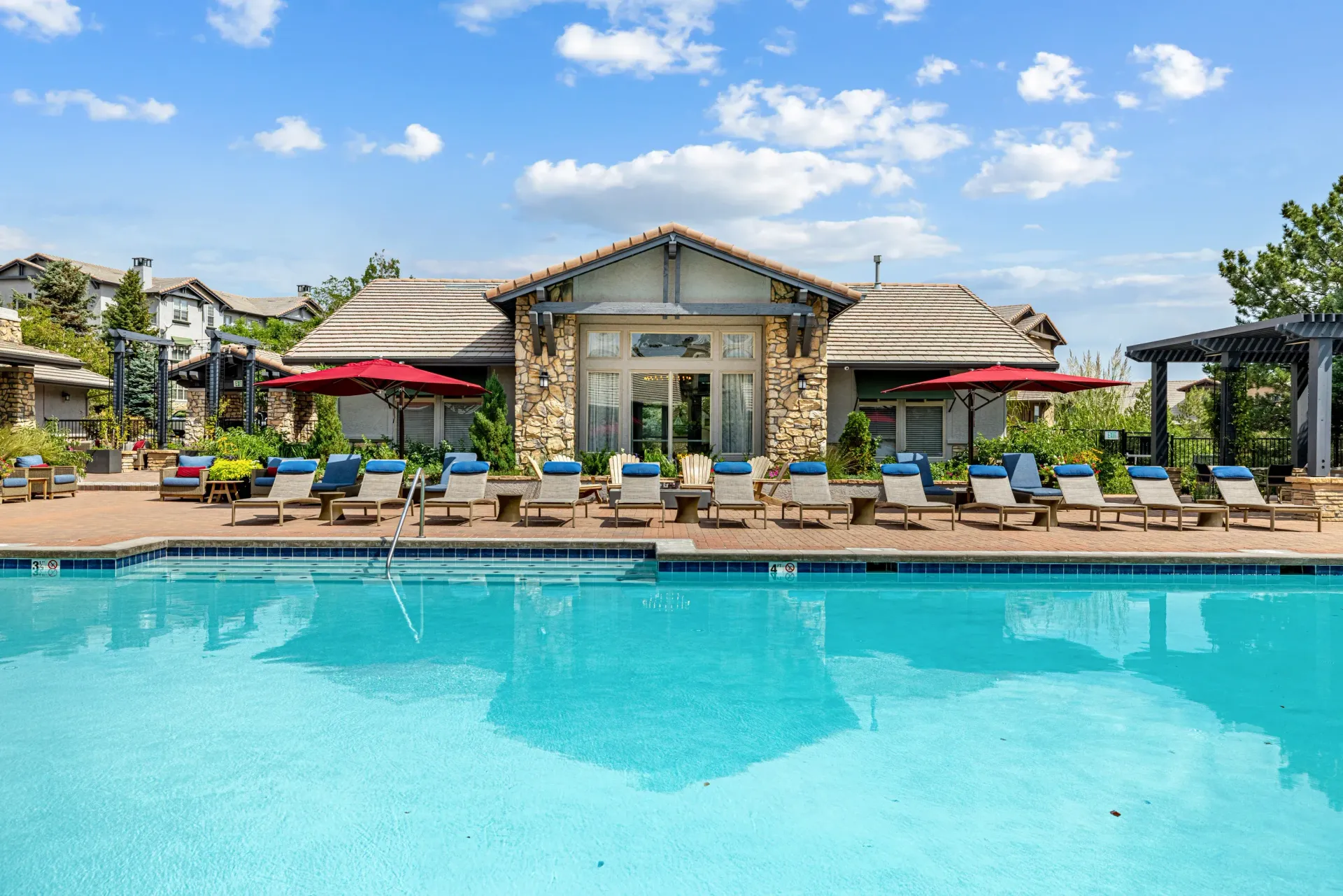 Outdoor pool with lounge chairs and red umbrellas by a clubhouse.