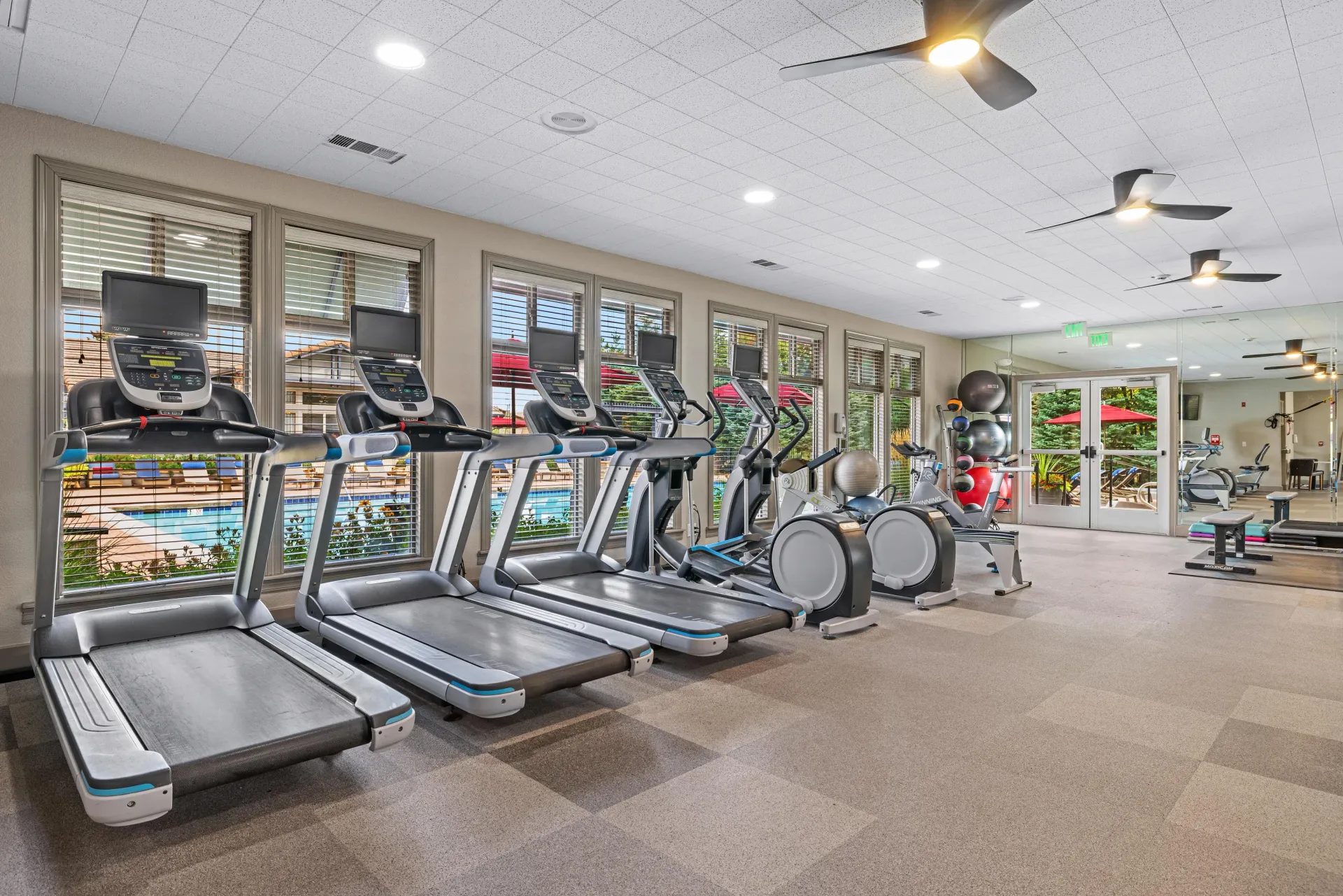 Indoor apartment gym with treadmills and cardio machines; windows overlook the outdoor pool.