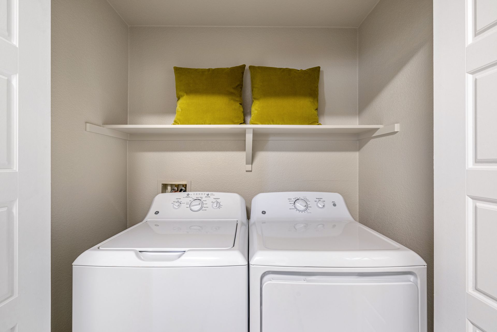 Laundry room with white washer and dryer, shelf with two yellow pillows.