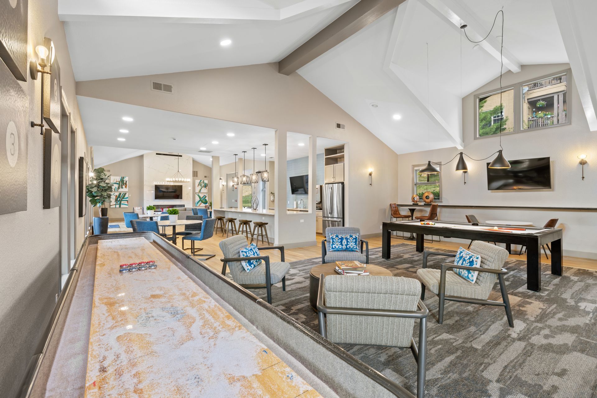 Lounge area with shuffleboard, seating, and open kitchen. Neutral tones, high ceiling, and natural light.