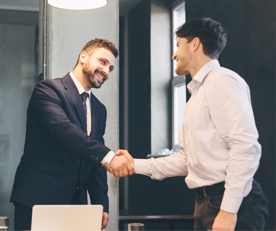 Two men in suits shaking hands, smiling, in an office.