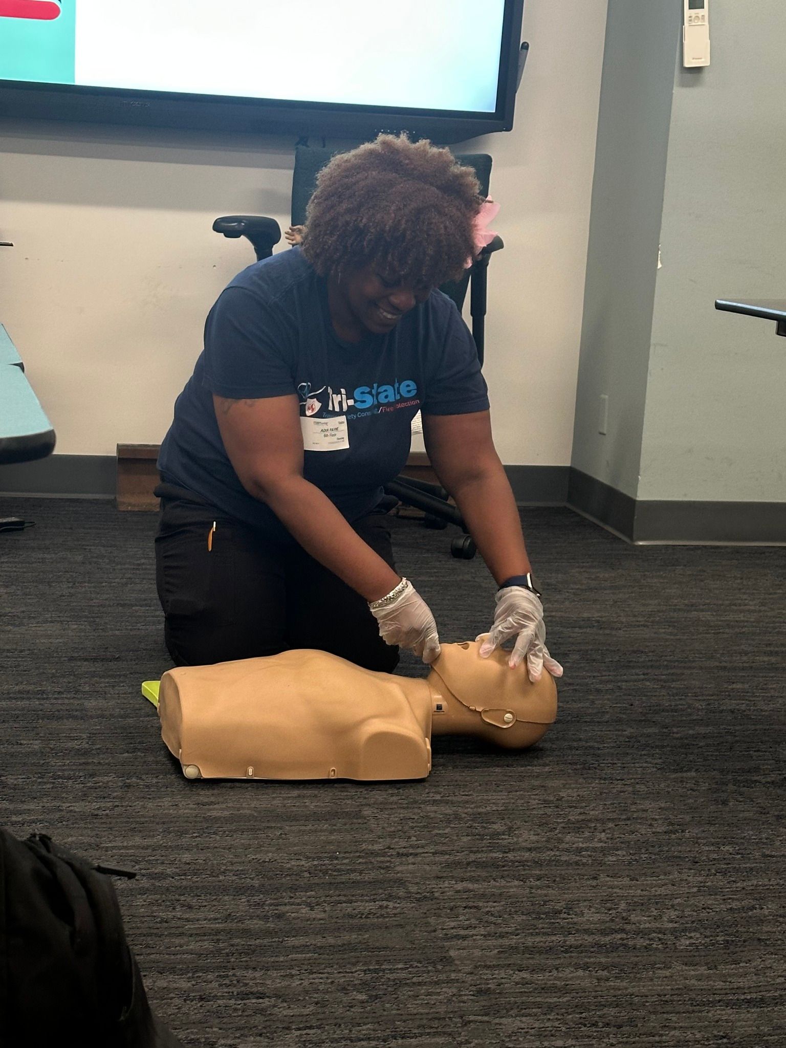 A Tri-State Training instructor kneels on carpet performing CPR training on a manikin in a classroom