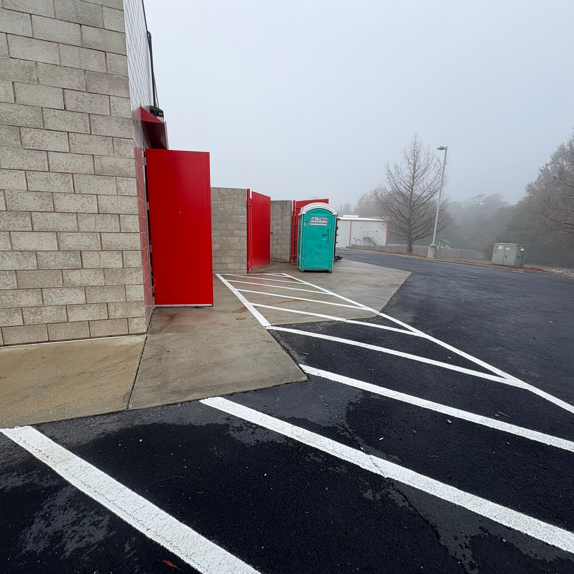 Foggy parking lot beside a stone building, with red doors, a portable toilet, and empty marked spaces.