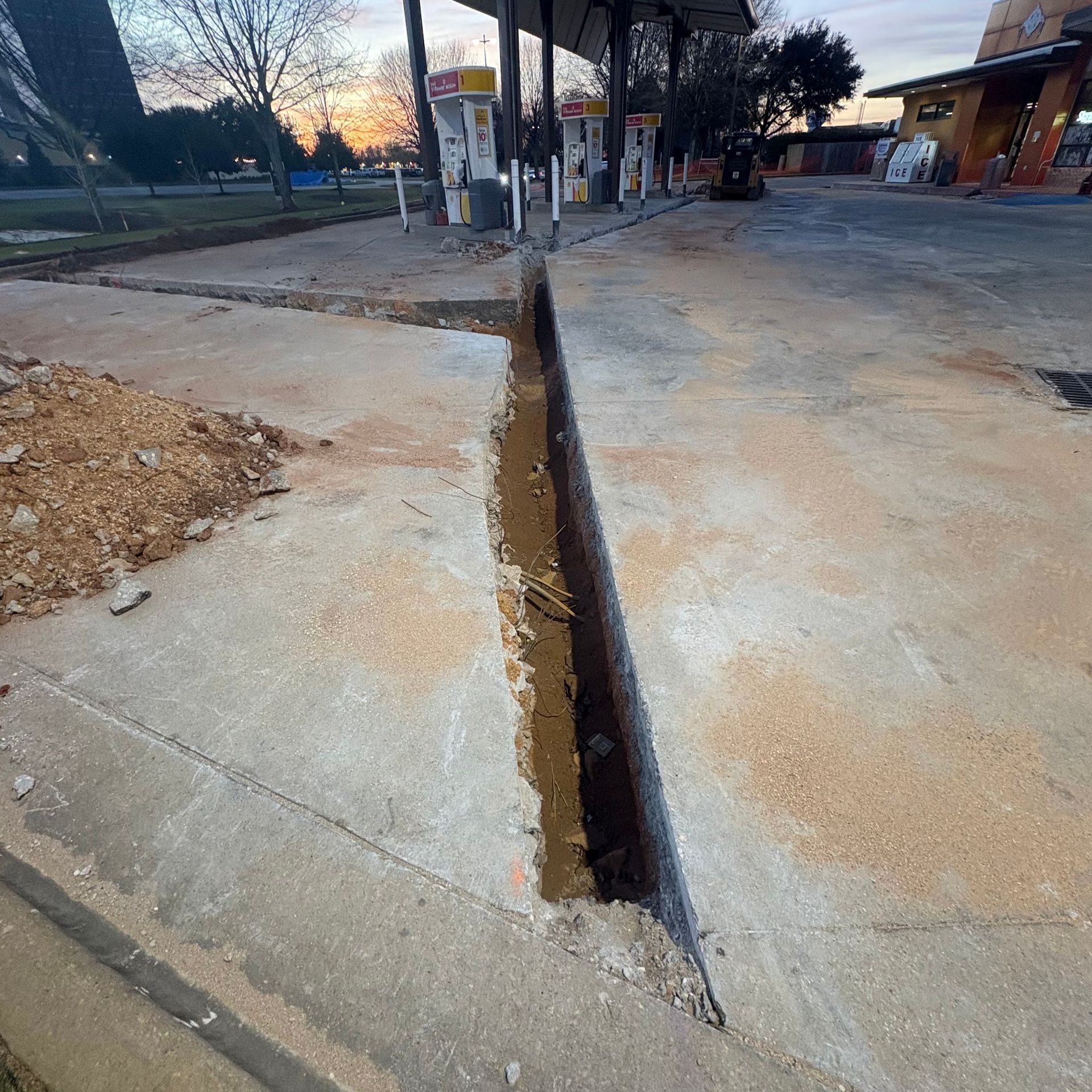 Wide concrete crack or trench running through a sidewalk near a gas station at dusk