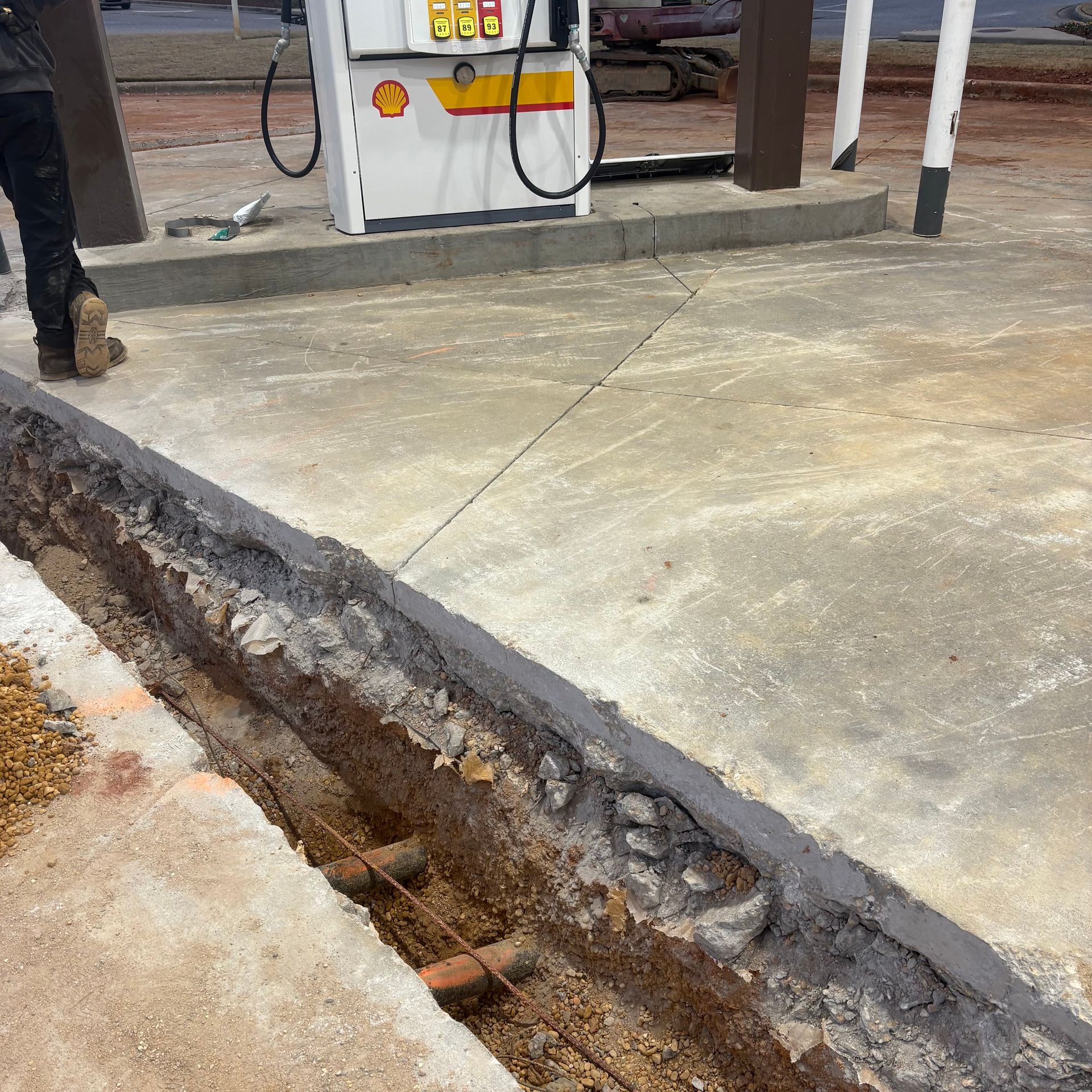 Cracked concrete trench beside a gas pump island with a worker’s legs visible on the left