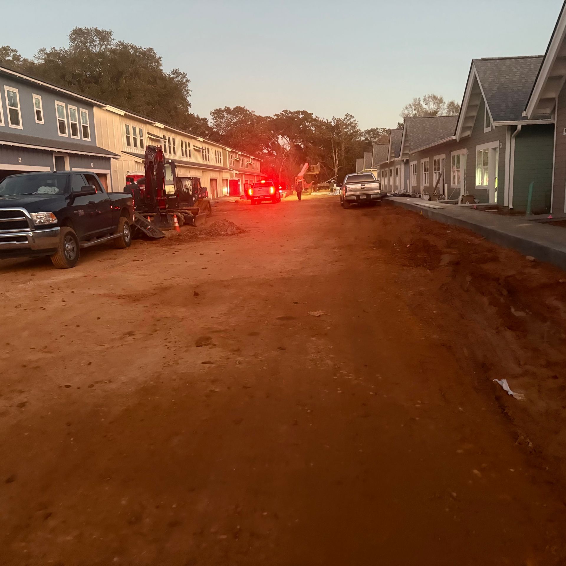 Dirt road in a residential area at dusk with parked cars and bright red emergency lights in the distance