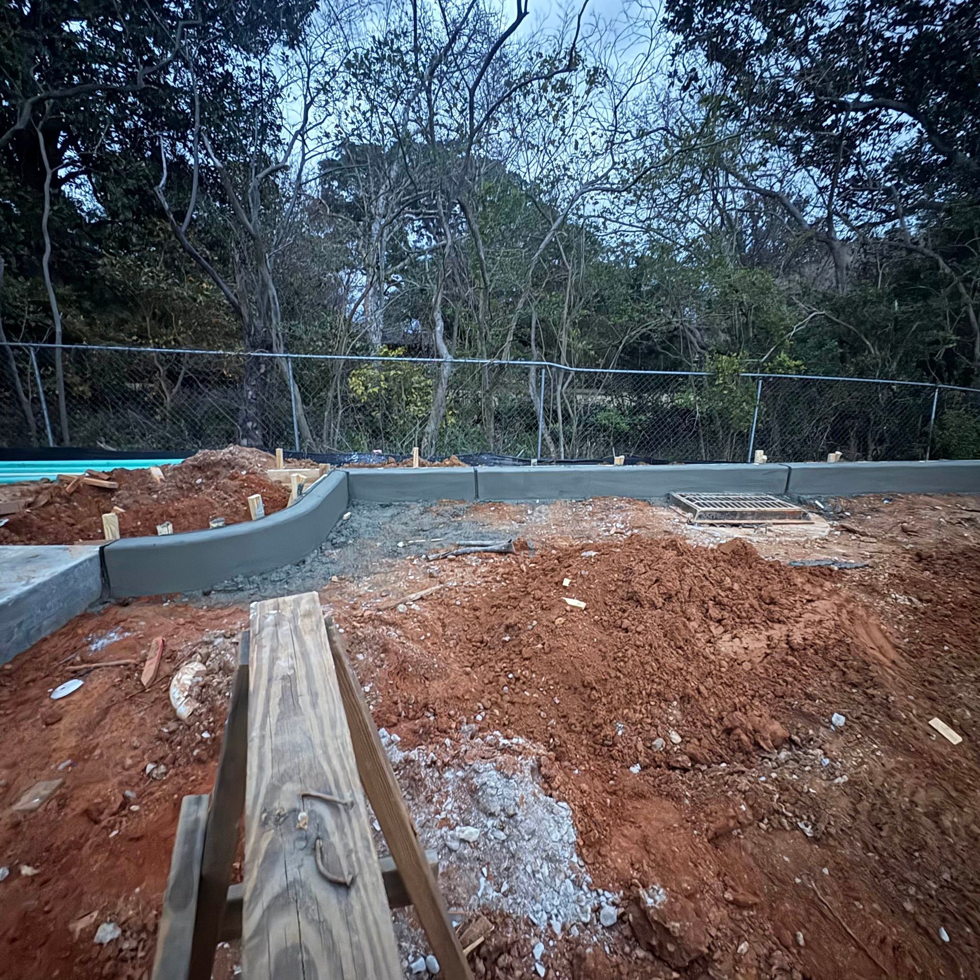 Curved concrete foundation wall beside red dirt construction site, with trees in the background