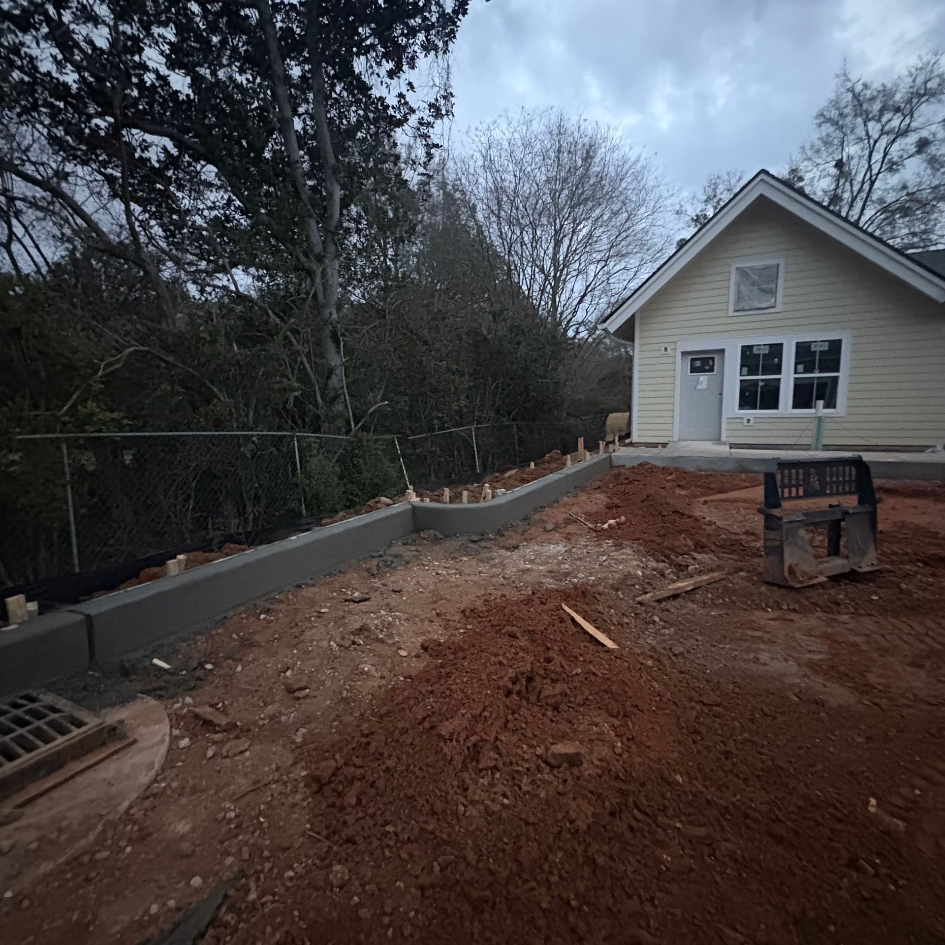 House with a dirt driveway and newly built retaining wall beside a chain-link fence at dusk