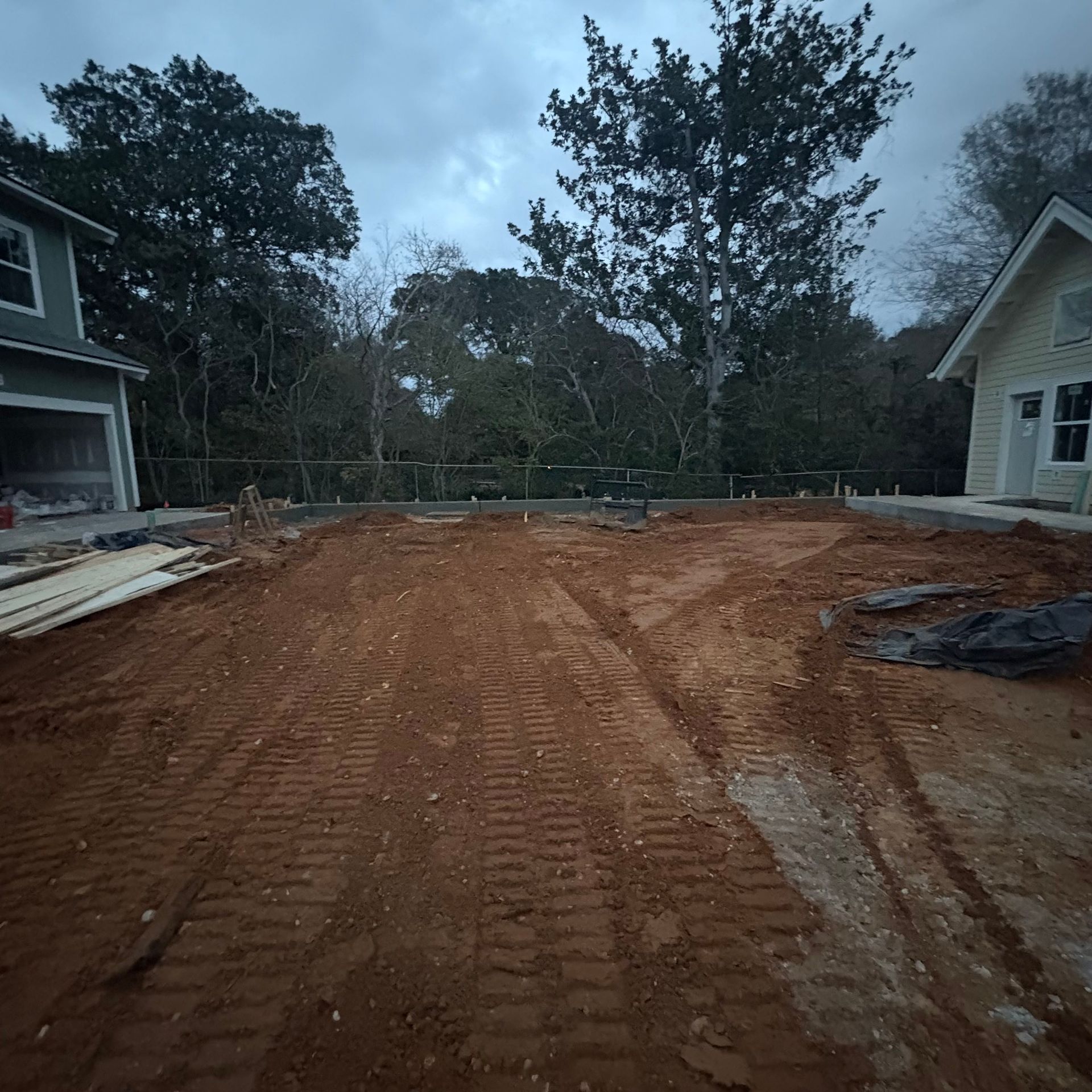 Freshly graded dirt lot between two houses at dusk, with tire tracks and trees in the background