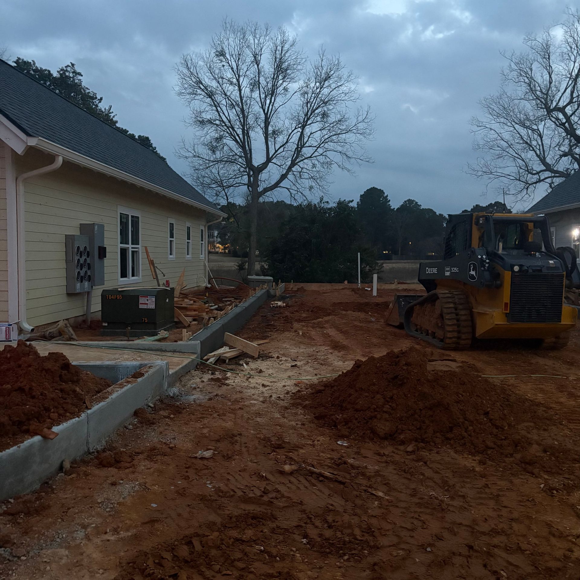 Construction site beside a house at dusk, with excavator and muddy ground work underway.