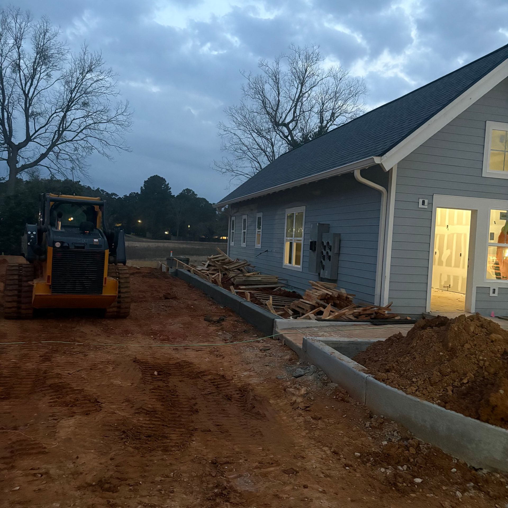 Construction site beside a gray house at dusk, with a yellow bulldozer and dirt piles along a driveway.