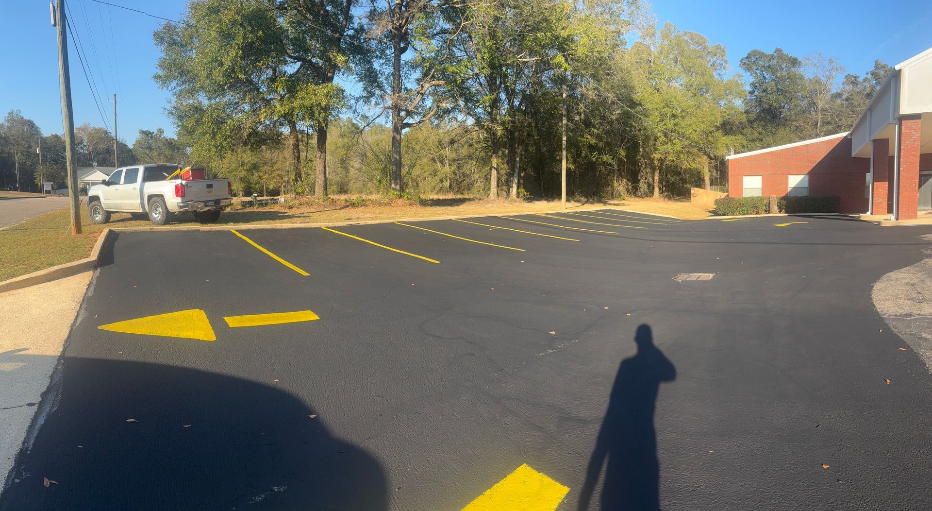 Empty parking lot with yellow directional arrows, trees in back, and a brick building on the right.