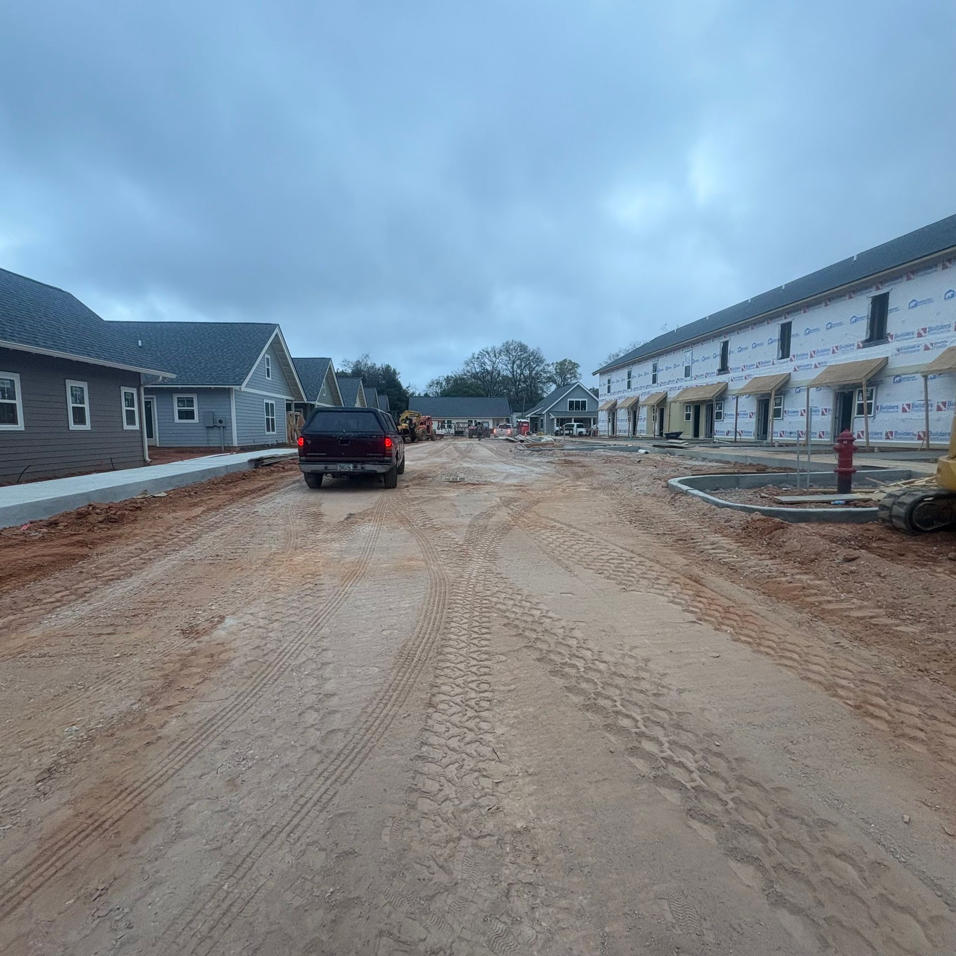 Dirt road under construction between two rows of houses, with a black SUV and cloudy sky.