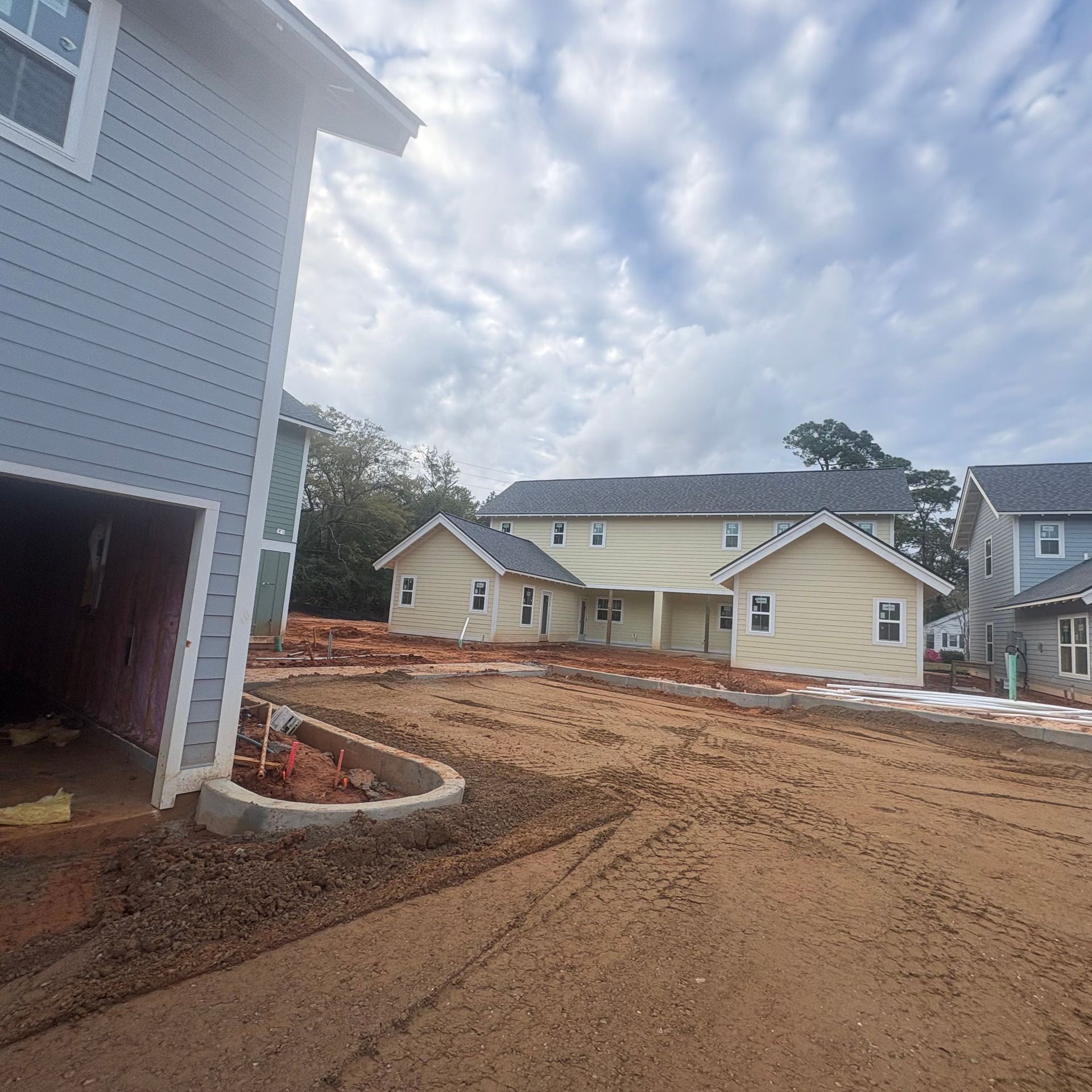 New homes under construction beside a dirt road, with a white house in the foreground and cloudy sky.