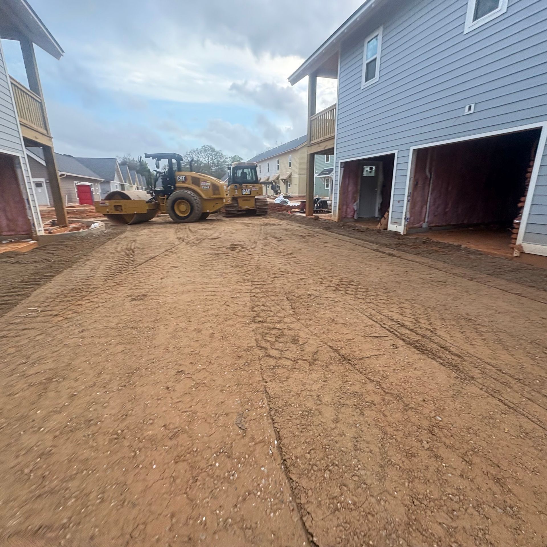 Construction site with a yellow loader grading a muddy driveway between houses.