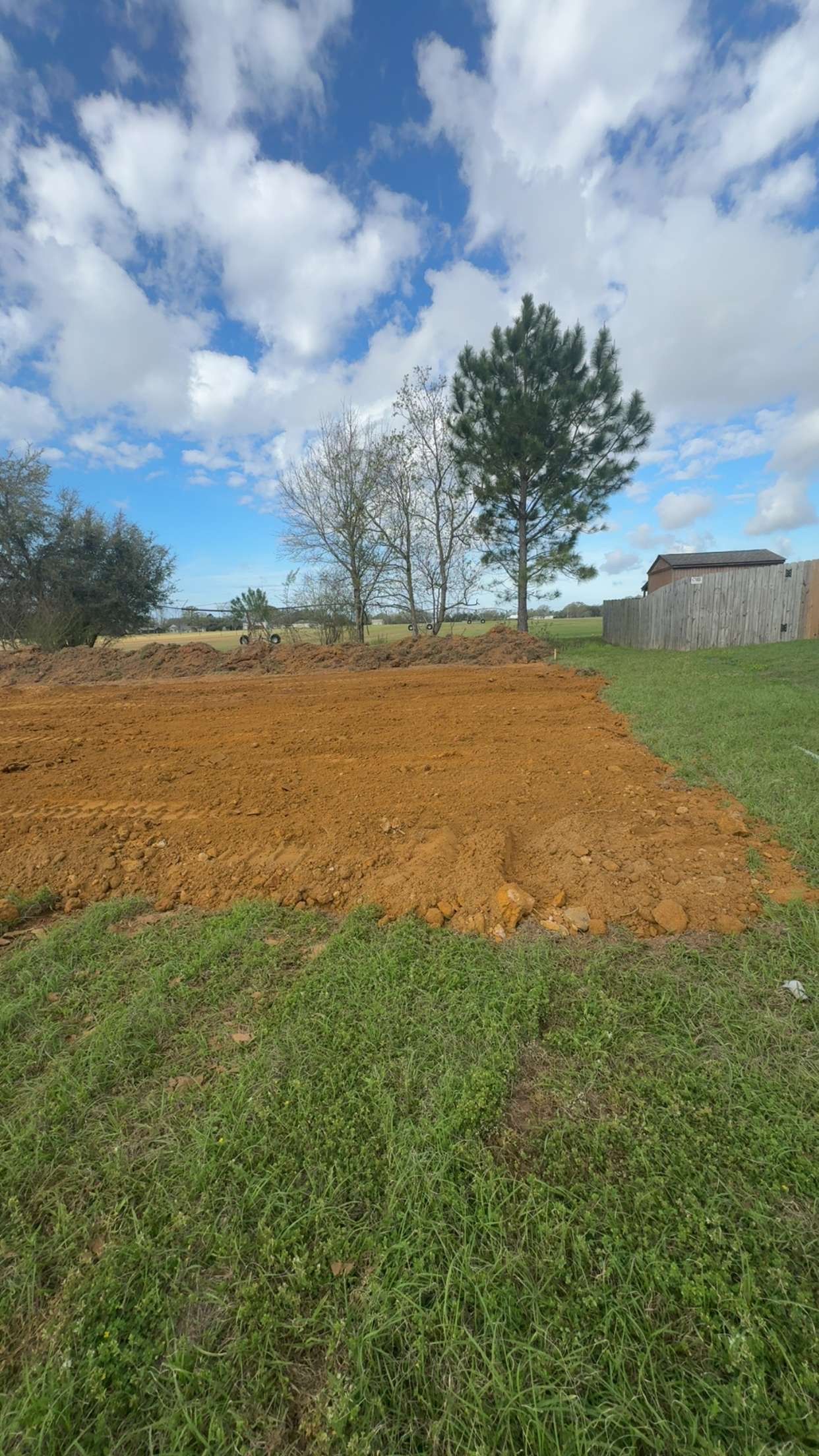 Freshly tilled brown field beside green grass under a cloudy blue sky with trees and a small shed