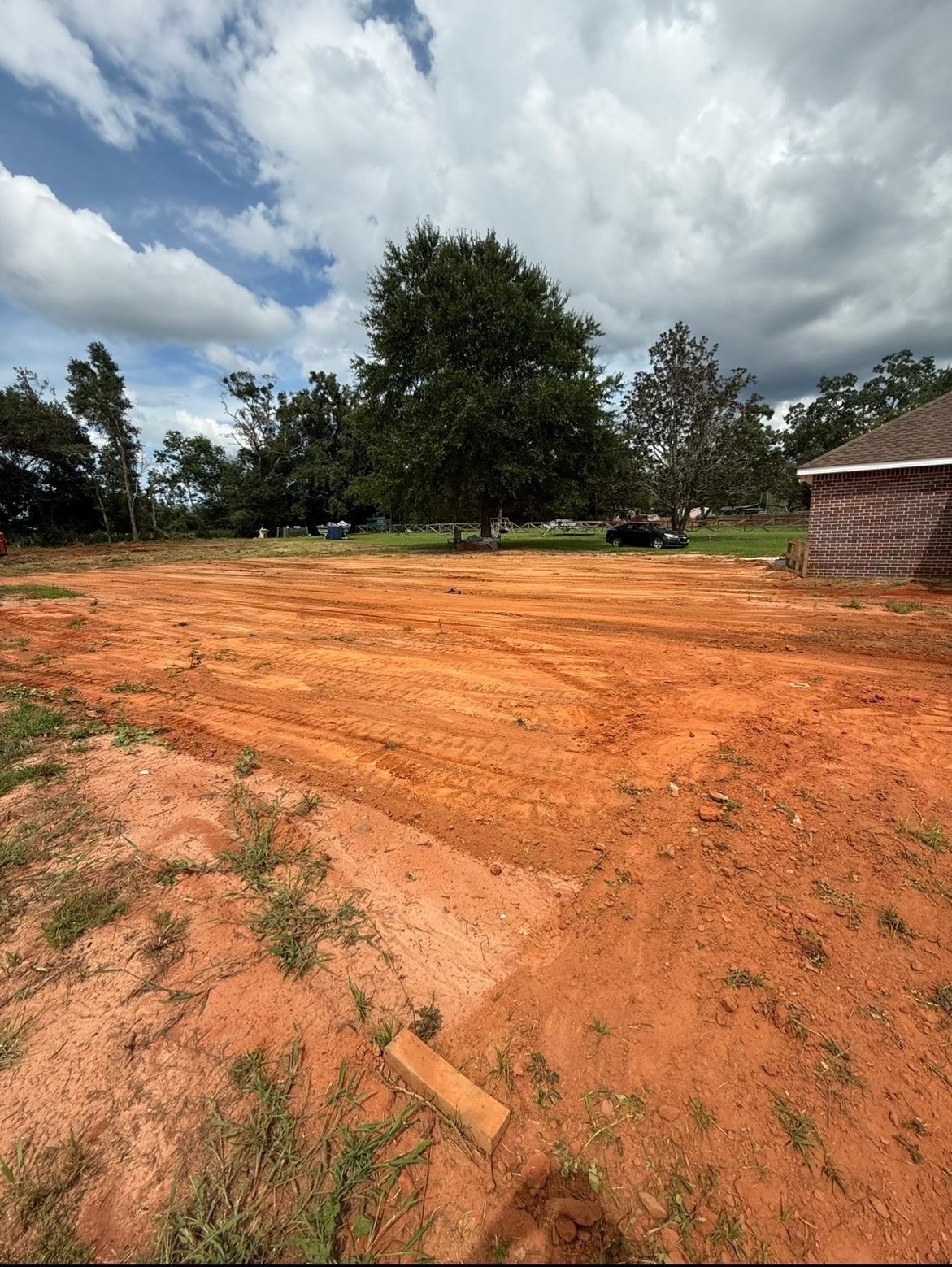 Dirt clearing with orange soil, trees, cloudy sky, and a house at the right edge.