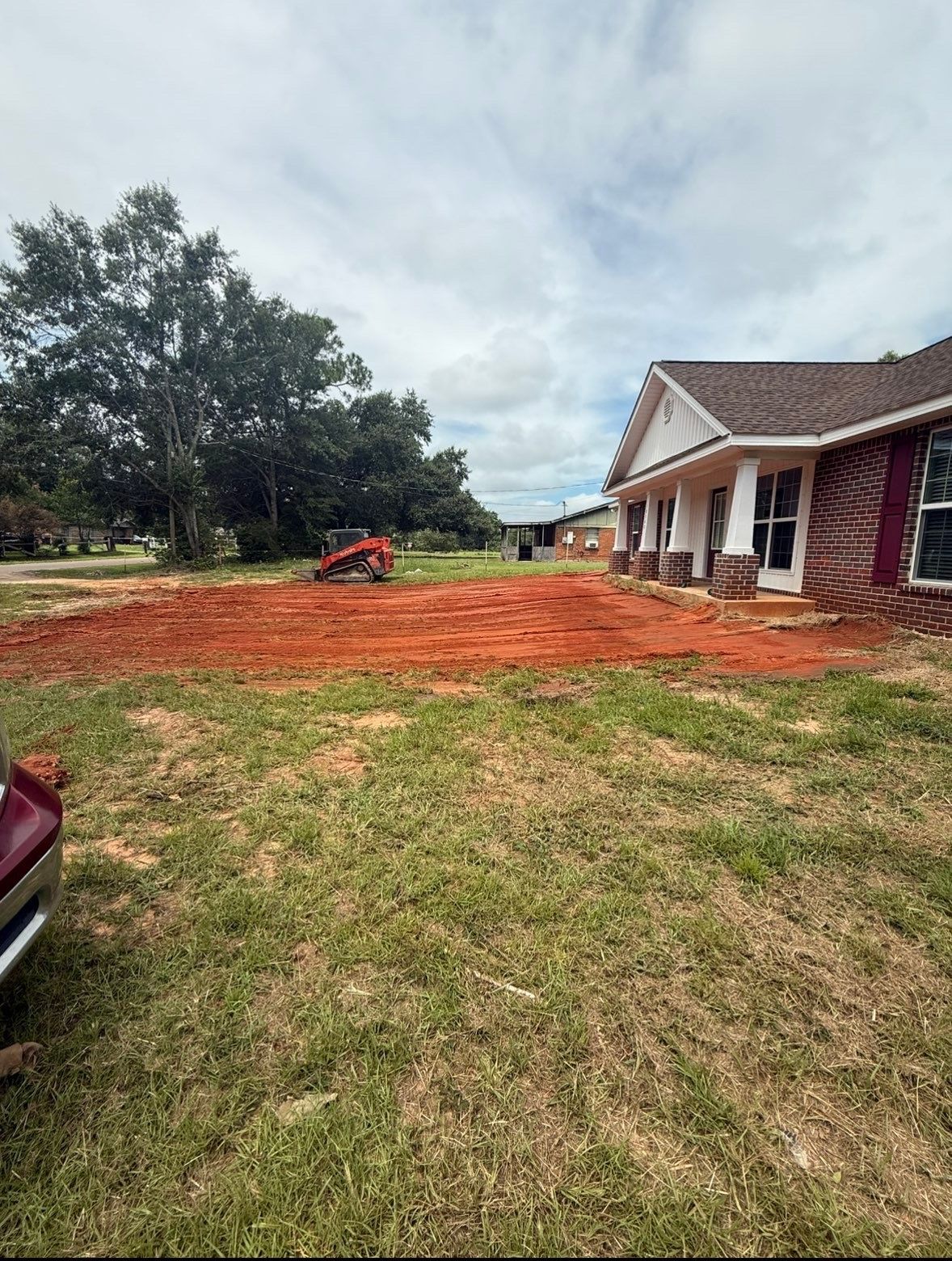Front yard with red dirt, a small excavator, and a brick house under a cloudy sky
