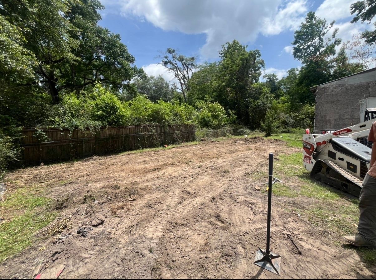 Cleared dirt lot beside trees and a fence, with a truck at right and a survey stake in the foreground