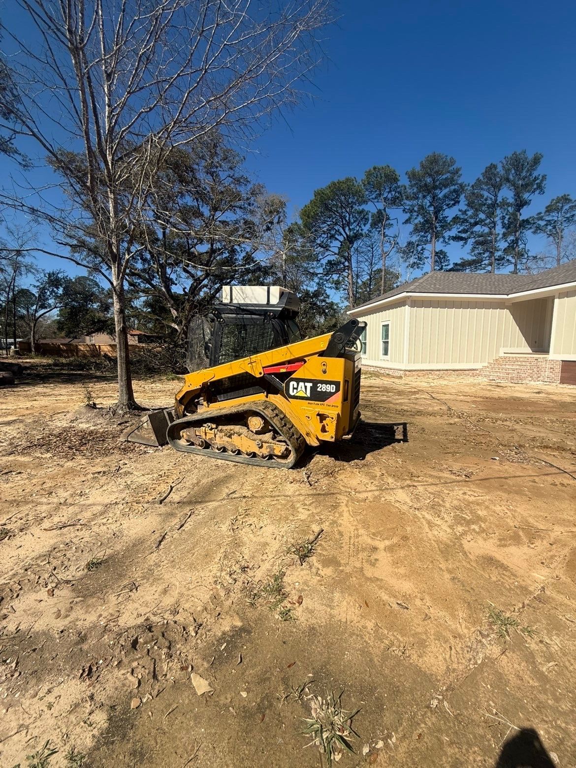 Yellow skid steer on dirt lot beside a beige mobile home, with trees and blue sky in the background