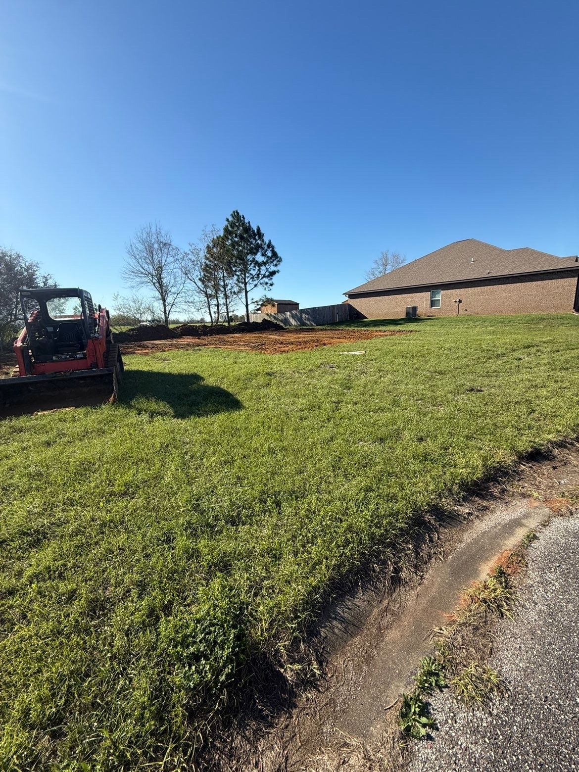Grassy hillside beside a brick house, with a red excavator and a clear blue sky.