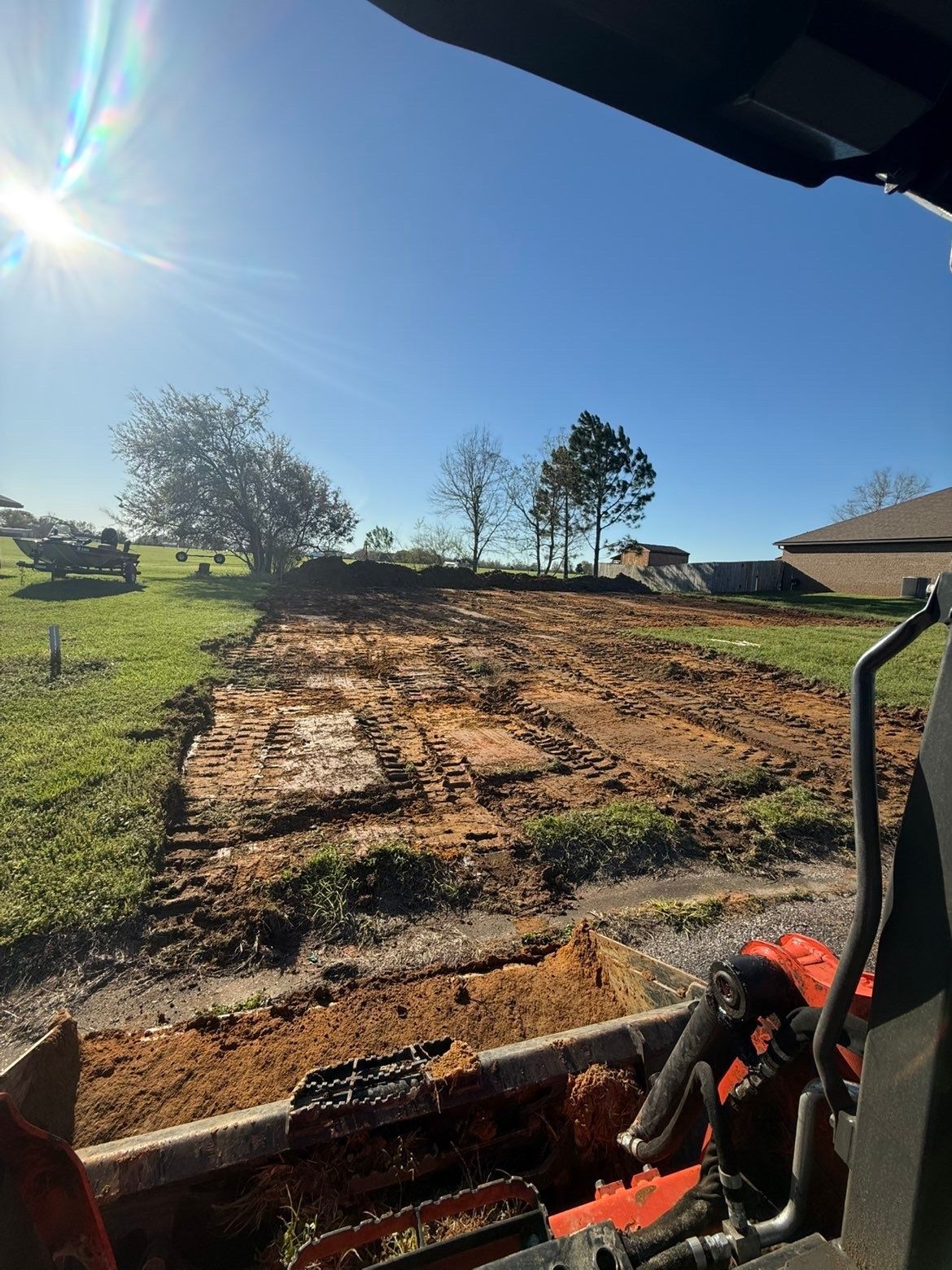 Excavator clearing a dirt lot under a bright blue sky, with trees and a house nearby.