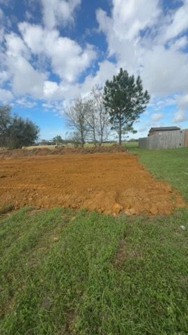 Freshly dug brown dirt patch in a grassy field under a cloudy blue sky, with trees and a small building nearby