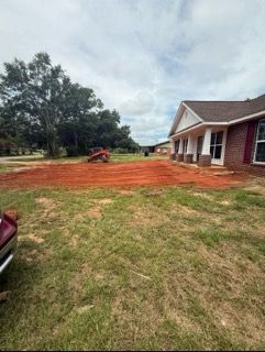 House with red dirt yard, orange excavator, and trees under a cloudy sky